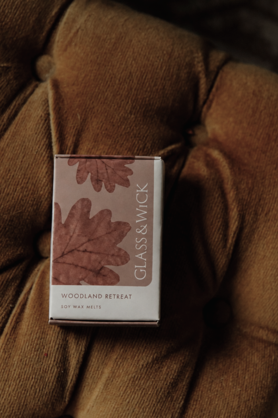 A rectangular box of GLASS & WICK Woodland Retreat soy wax melts sits on a brown tufted velvet surface. The box features a dusty pink panel with brown oak leaf illustrations and white text.
