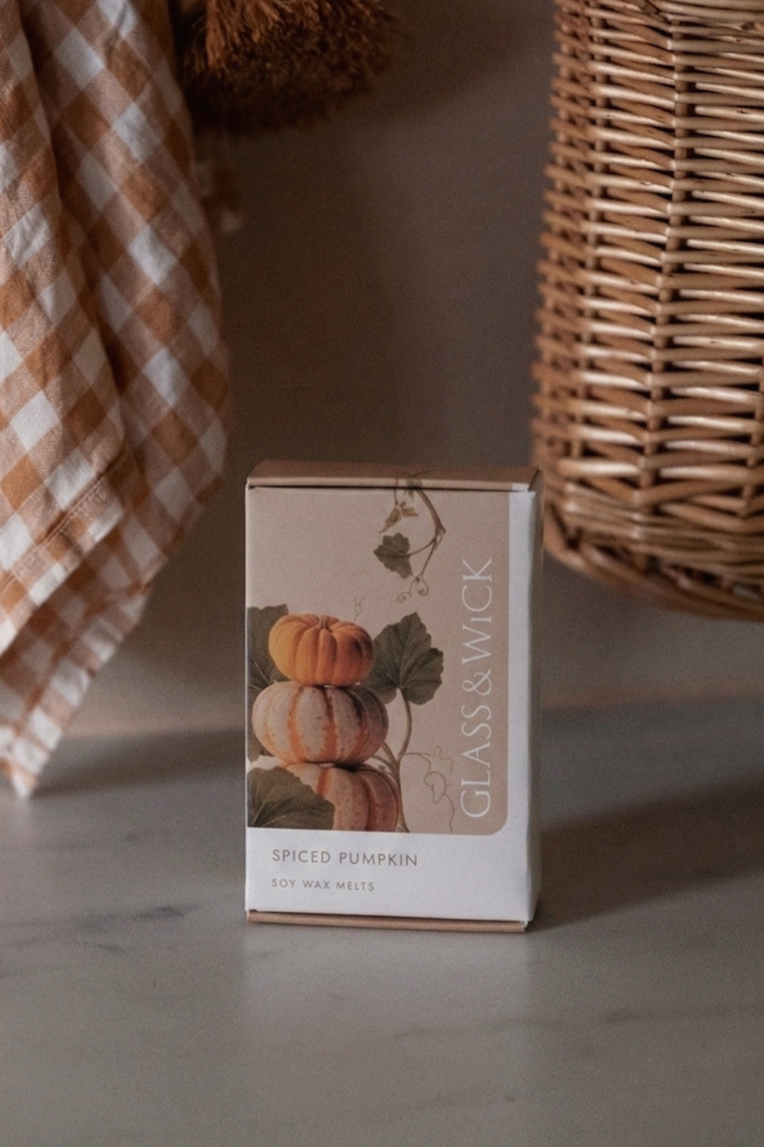 6 pumpkin wax melts GLASS&WICK SPICED PUMPKIN sits on a countertop, with a brown-and-white gingham cloth on the left and a woven basket on the right in a softly lit indoor setting.