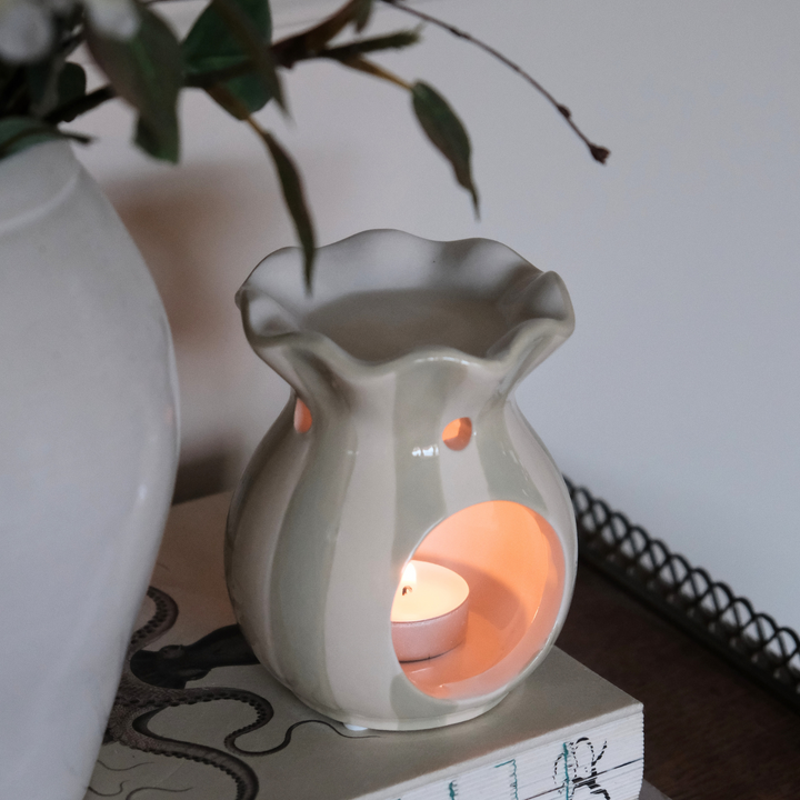 White wax warmer with a lit candle inside on a surface with a plant in the background