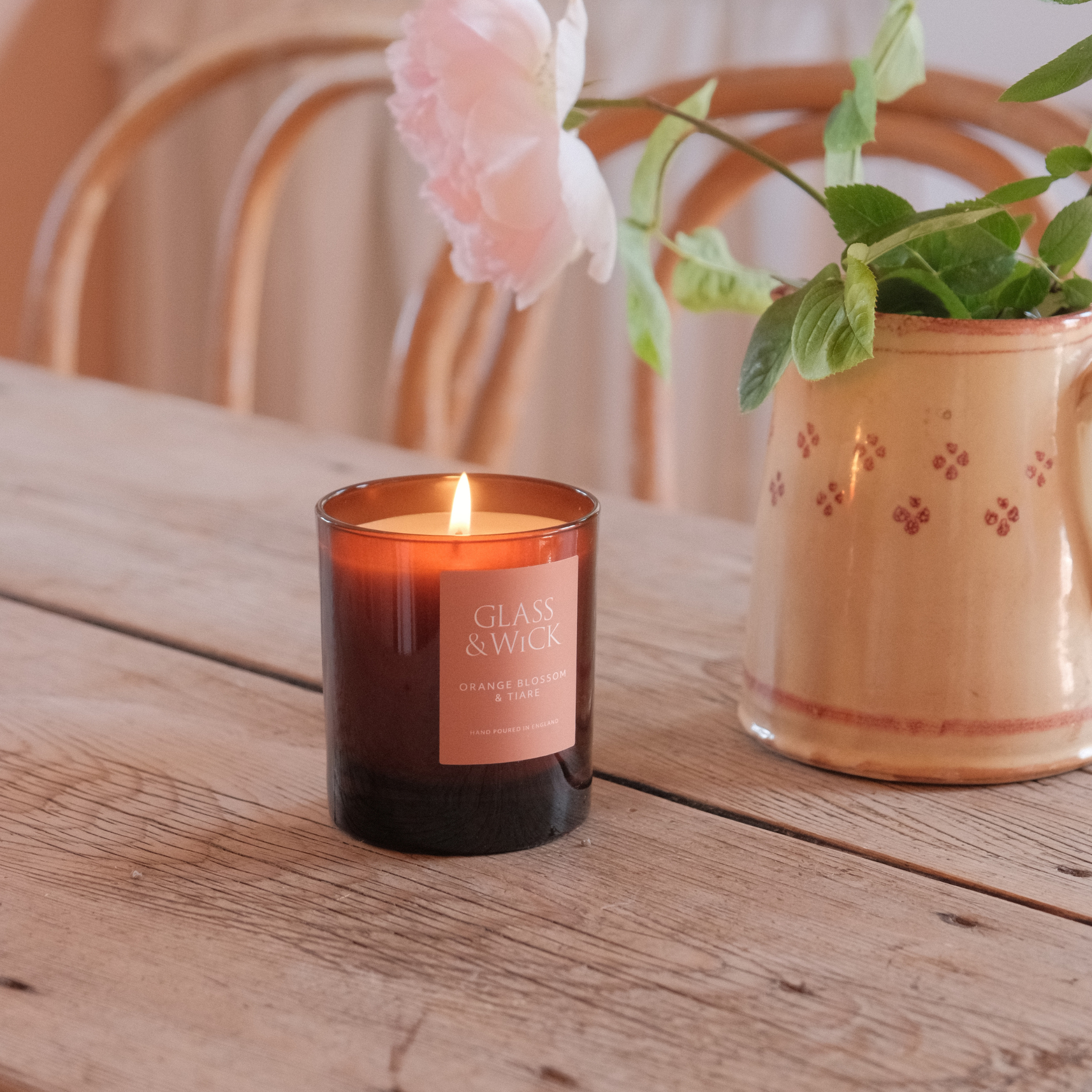 Glass & Wick candle on a wooden table with a vase of flowers in the background