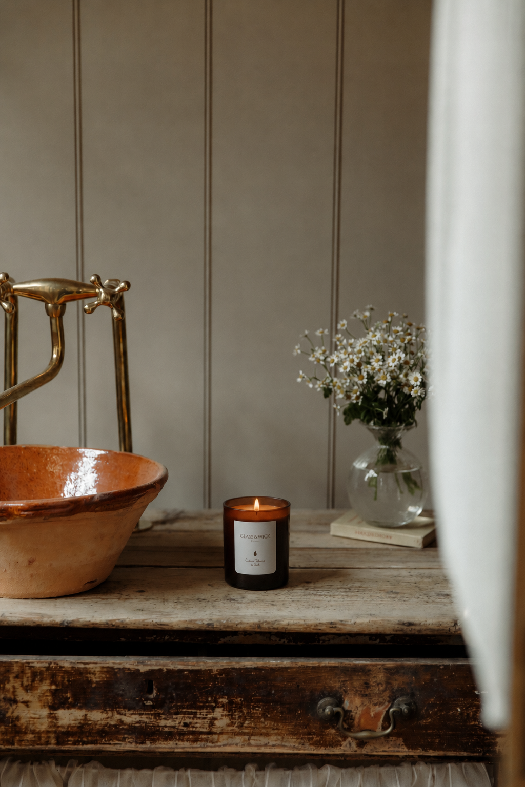 A lit amber glass candle sits on a rustic wooden table next to a terracotta bowl and a vase of small white flowers, set against a beige paneled wall.